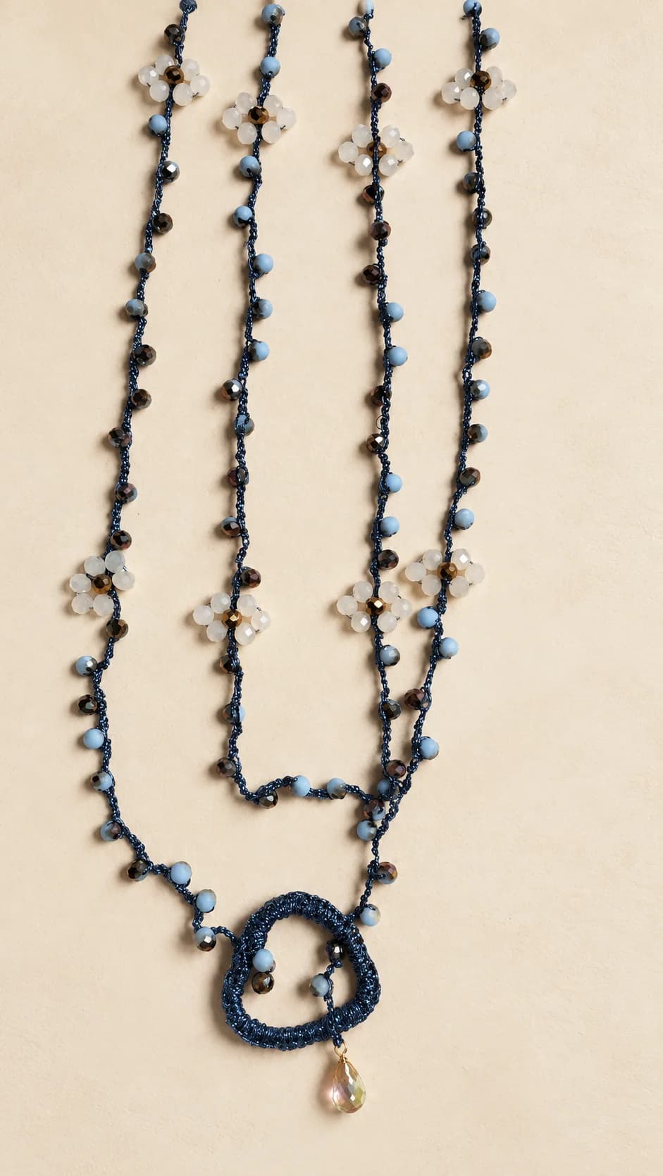 Fili con piccoli elementi blu e fiori chiari; pendente a cuore dal bordo blu con una piccola goccia chiara.