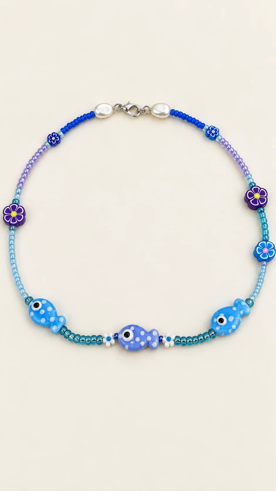 Collana con tre elementi a forma di pesce azzurri e lavanda, piccoli fiori blu e viola e perline coordinate. La foto originale comprende altri gioielli e un pesce verde sciolto, non inclusi.